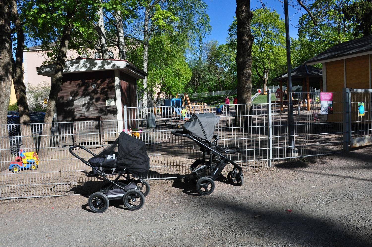 Två barnvagnar står parkerade utanför en inhägnad lekpark.