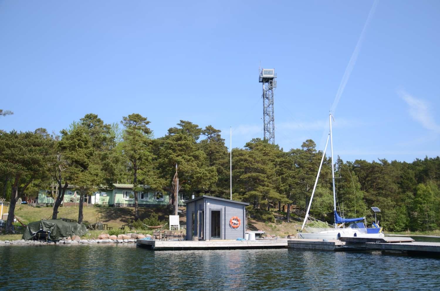 En lugn vik med en brygga, en segelbåt och en bastu på en flotte. I bakgrunden ett vakttorn.