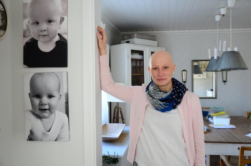 Ung kvinna utan hår tittar in i kameran. På väggen bredvid henne finns två babyporträtt, där barnen också är utan hår