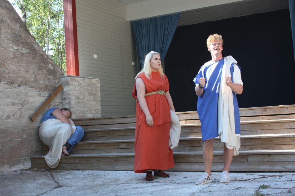 Alice Ekholm klädd i röd klänning och Jesper Lindblom klädd i blå dräkt står framför Kalkholmsestradens trappa. Fanny Stenström sitter på trappan bakom dem.