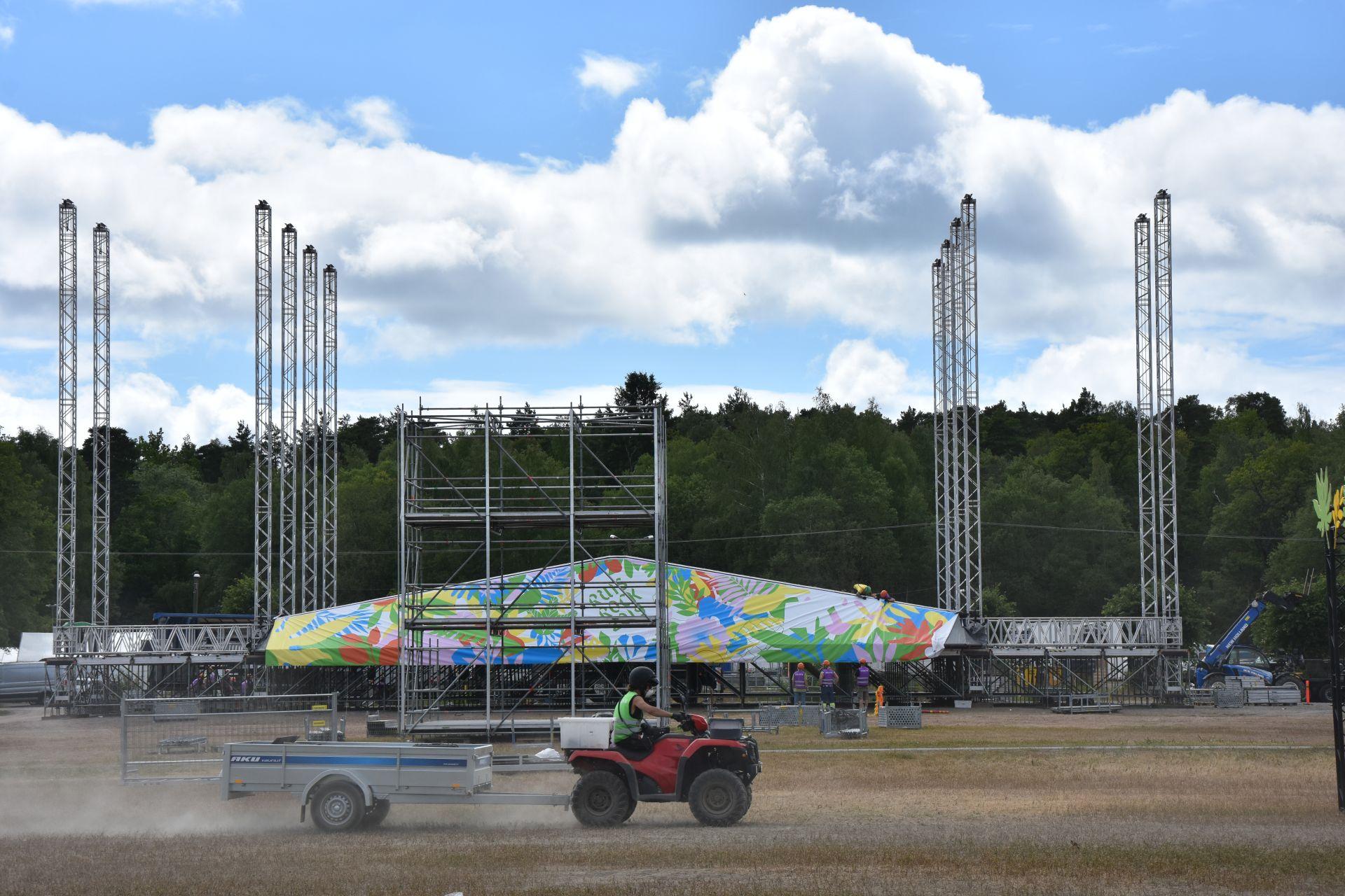 En festivalscen håller på att byggas. Scenen har fortfarande inget tak och framför scenen kör en person klädd i arbetskläder på en fyrhjuling.