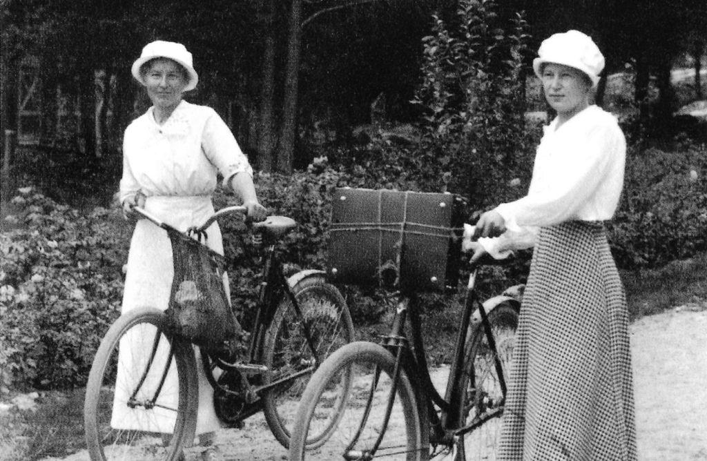Ett svartvitt fotografi av två kvinnor som leder varsin cykel.