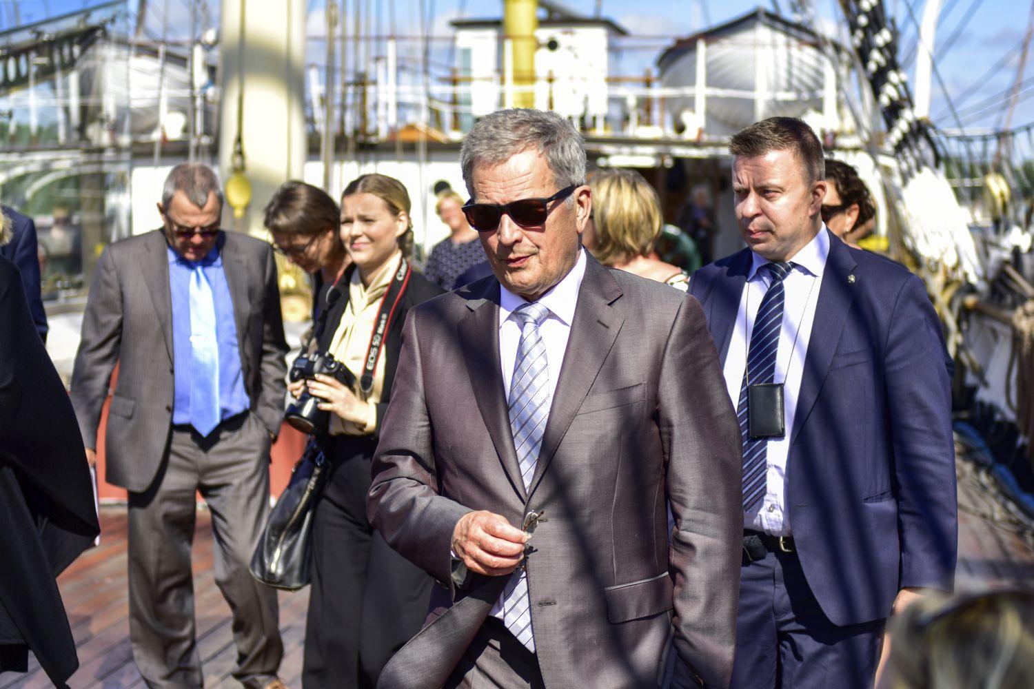 Sauli Niinistö iklädd kostym och solglasögon. I bakgrunden syns en hel del andra människor.