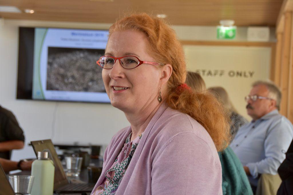 En kvinna står i ett konferensrum, i bakgrunden syns andra människor och på väggen projiceras en presentation.