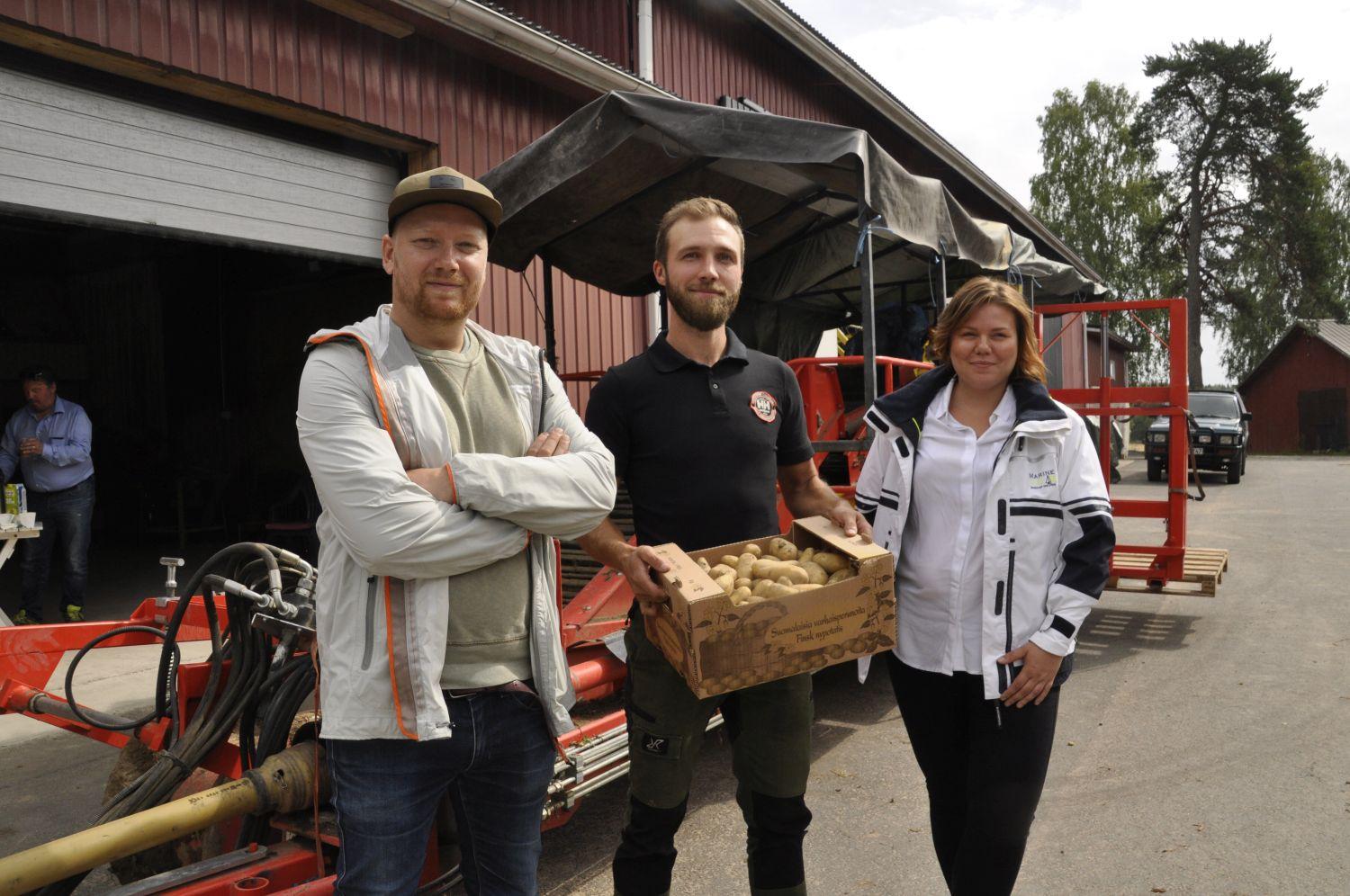 Två män och en kvinna står framför en potatisupptagare. mannen i mitten har en potatislåda i famnen.