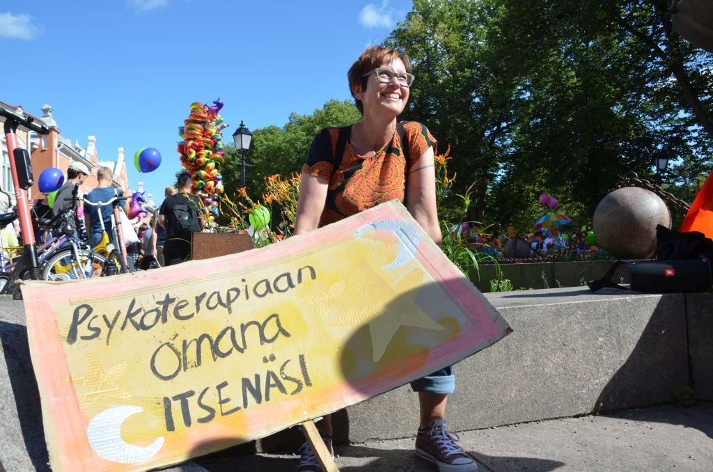 En kvinna sitter på kanten av en fonten och ler. Med sig har hon en skylt med texten "psykoterapiaan omana itsenä"