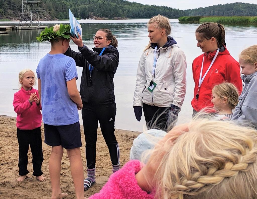 Barn och vuxna är samlade på en strand. En vuxen kvinna lägger en blomkrans på en pojkes huvud.