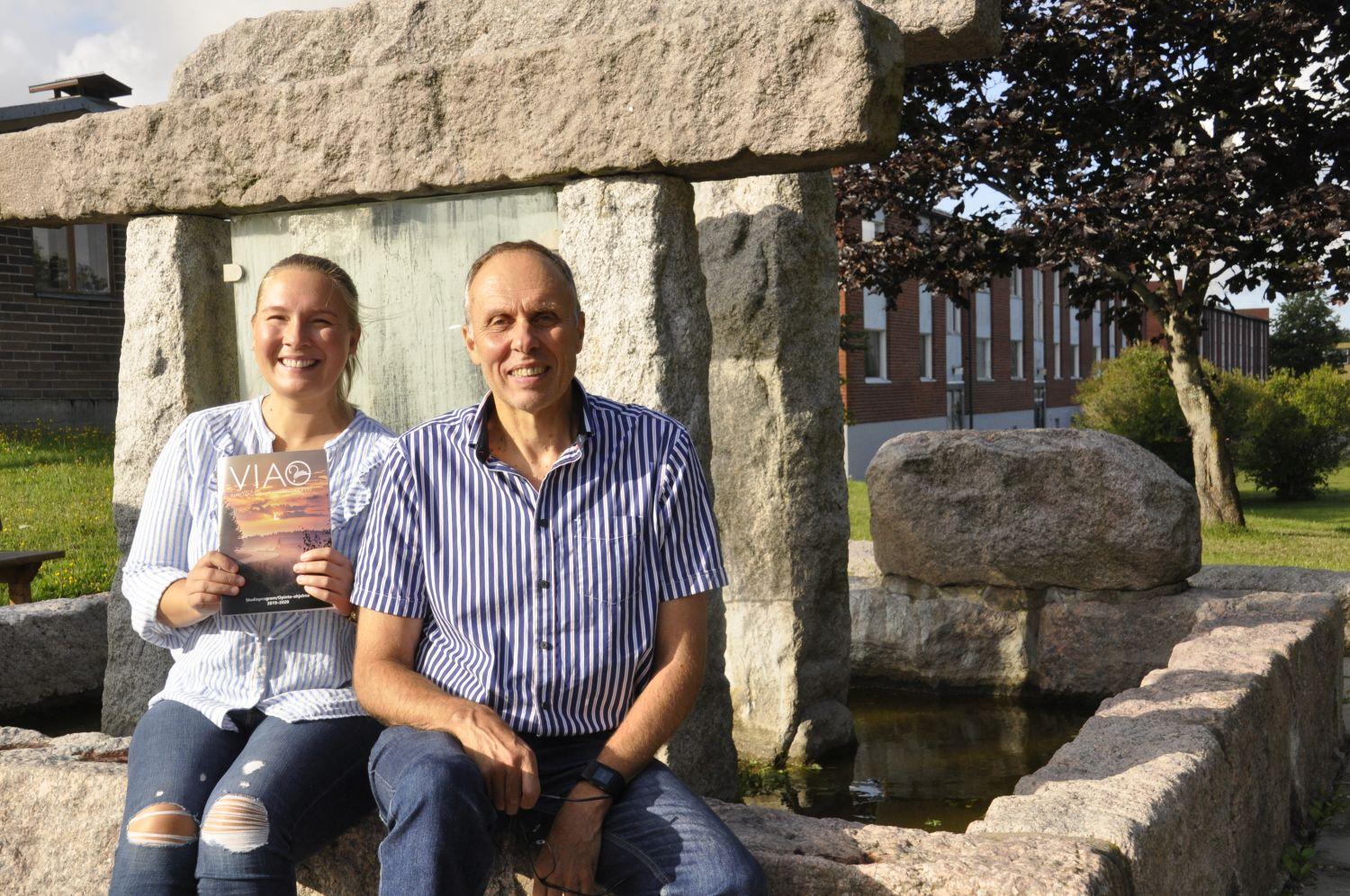 en kvinna och en man sitter vid ett monument i sten . Har en broschyr i handen.
