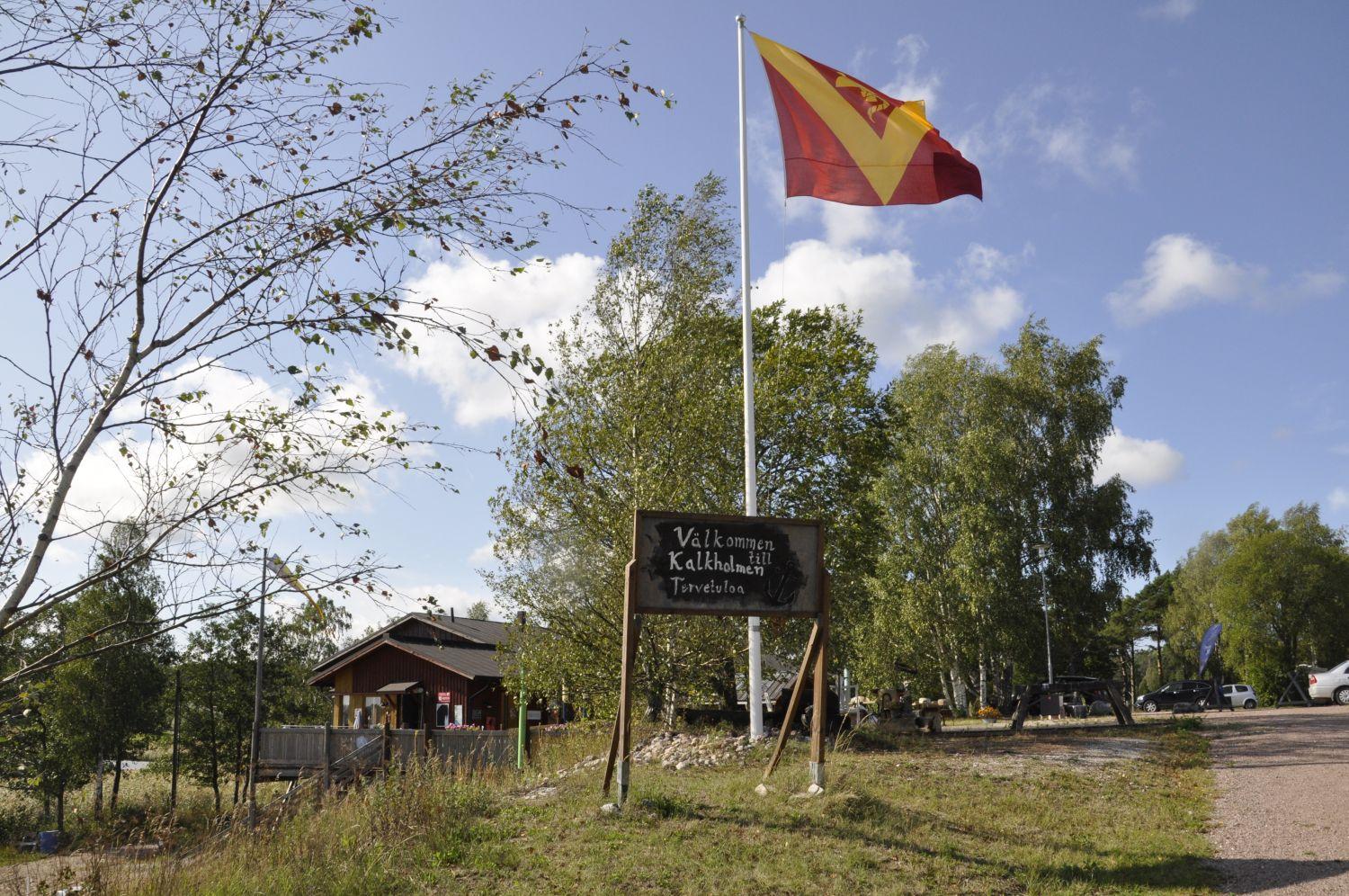 naturfint landskap med en byaflagga och en skylt: Välkommen till Kalkholmen. Foto: Emilia Örnmark.