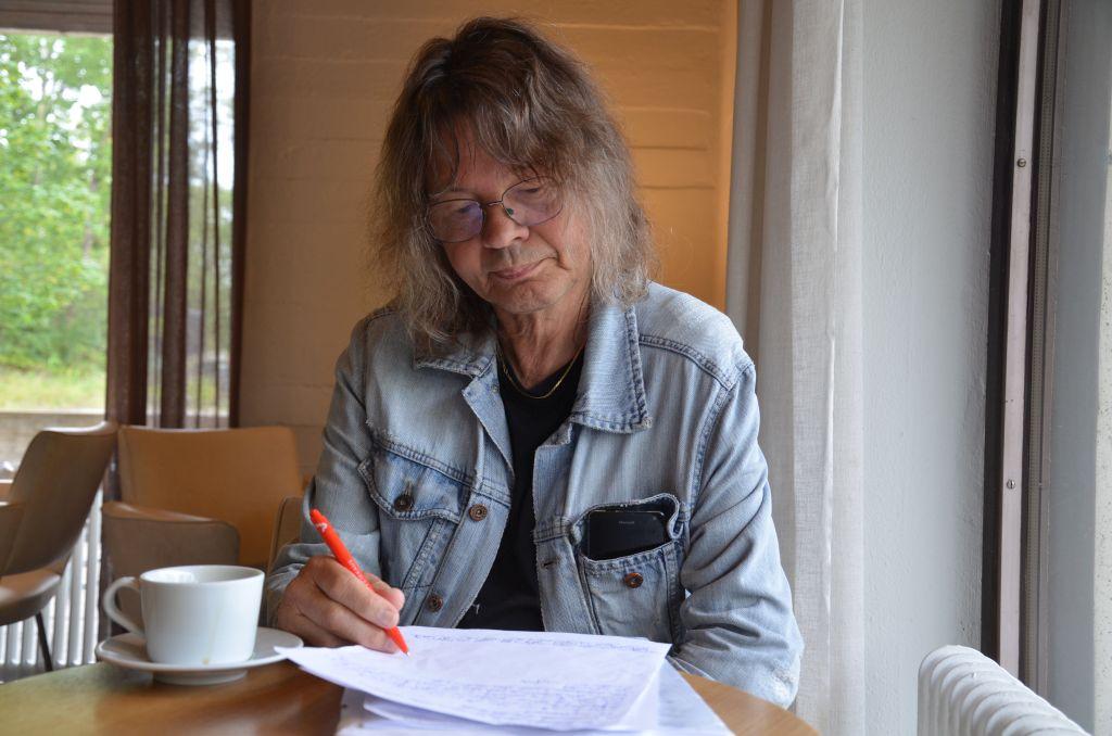 En långhårig man i jeansrock sitter på ett kafé och skriver. Framför sig har han et papper med anteckningar och ett kopp kaffe.