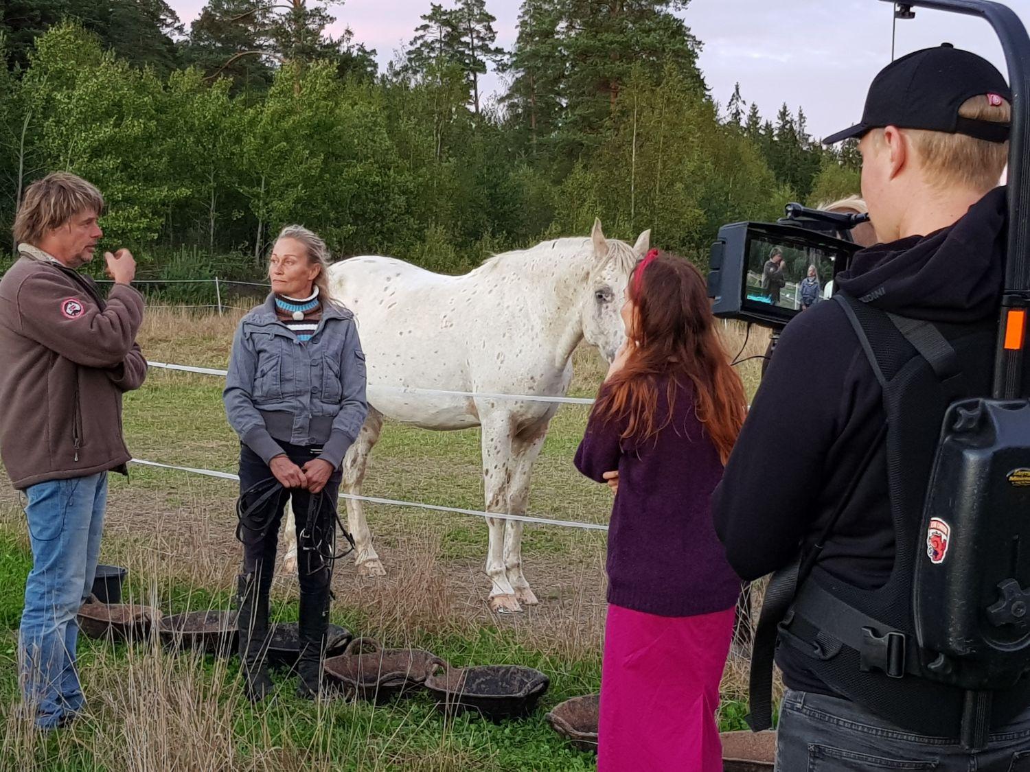 Ett filmteam bakifrån. Framför deras kamera syns en programledare, samt de två intervjuade framför en hästhage och en vit häst.