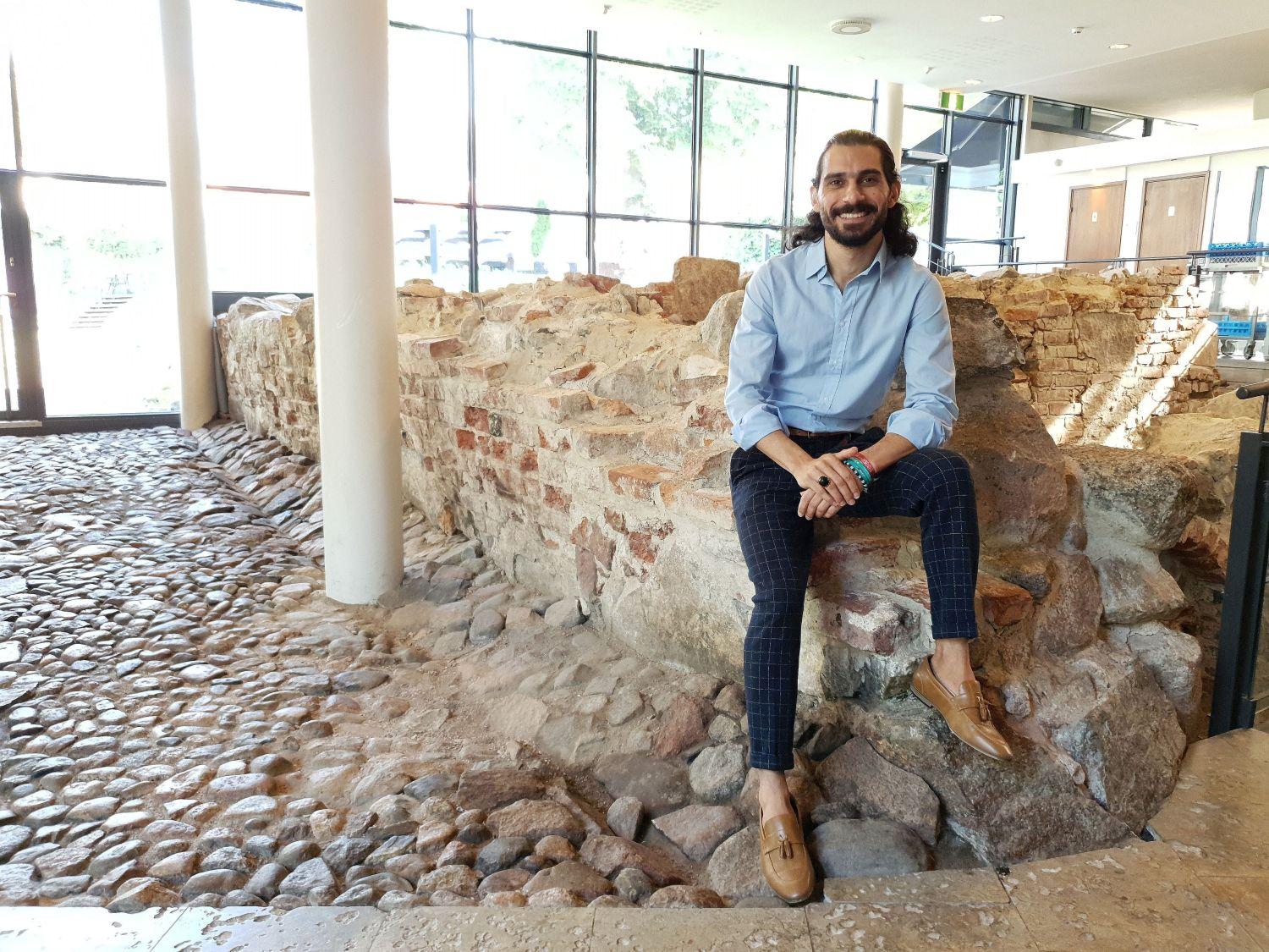 Bahaulddin Rawi sitter på ruinerna inne i Aboa Vetus café och ler mot kameran. Han är iklädd blå skjorta och mörka, rutiga byxor.