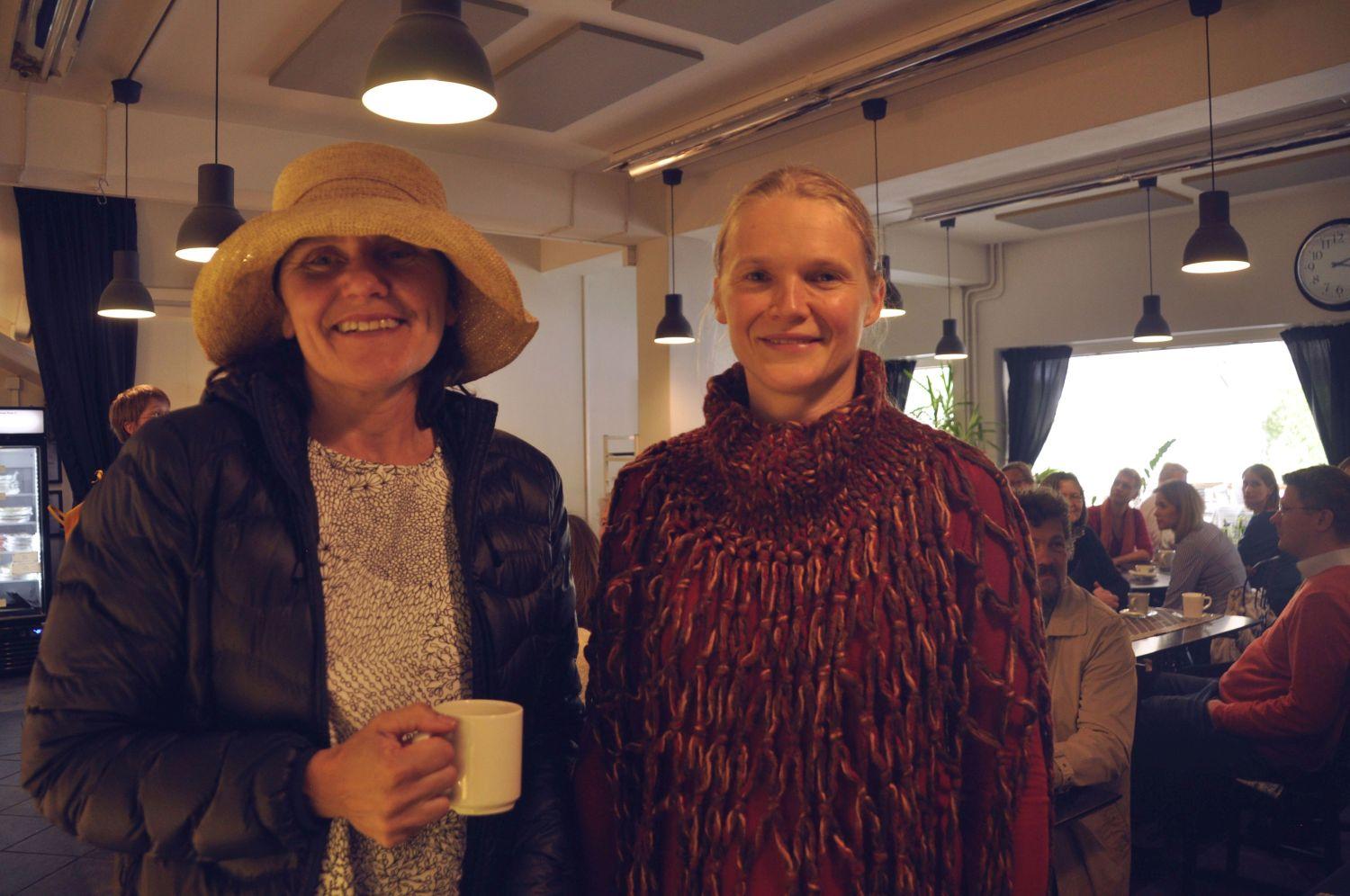 Två glada damer. I bakgrunden ett café fullt av folk.