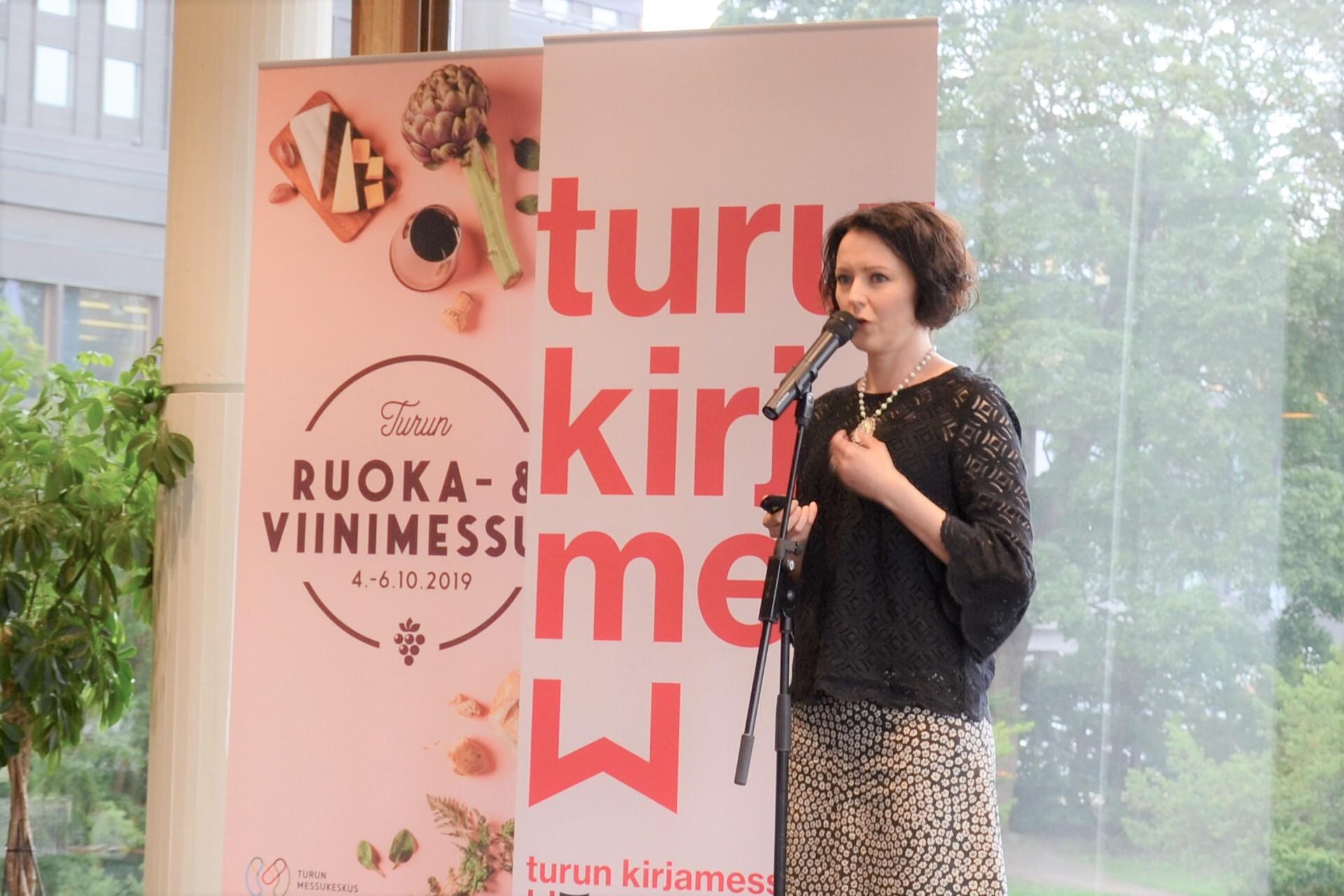Jenny Haukio, iförd svart blus och rutig kjol, står med micken i hand framför två roll ups med reklam för bokmässan och mat- och vinmässan.