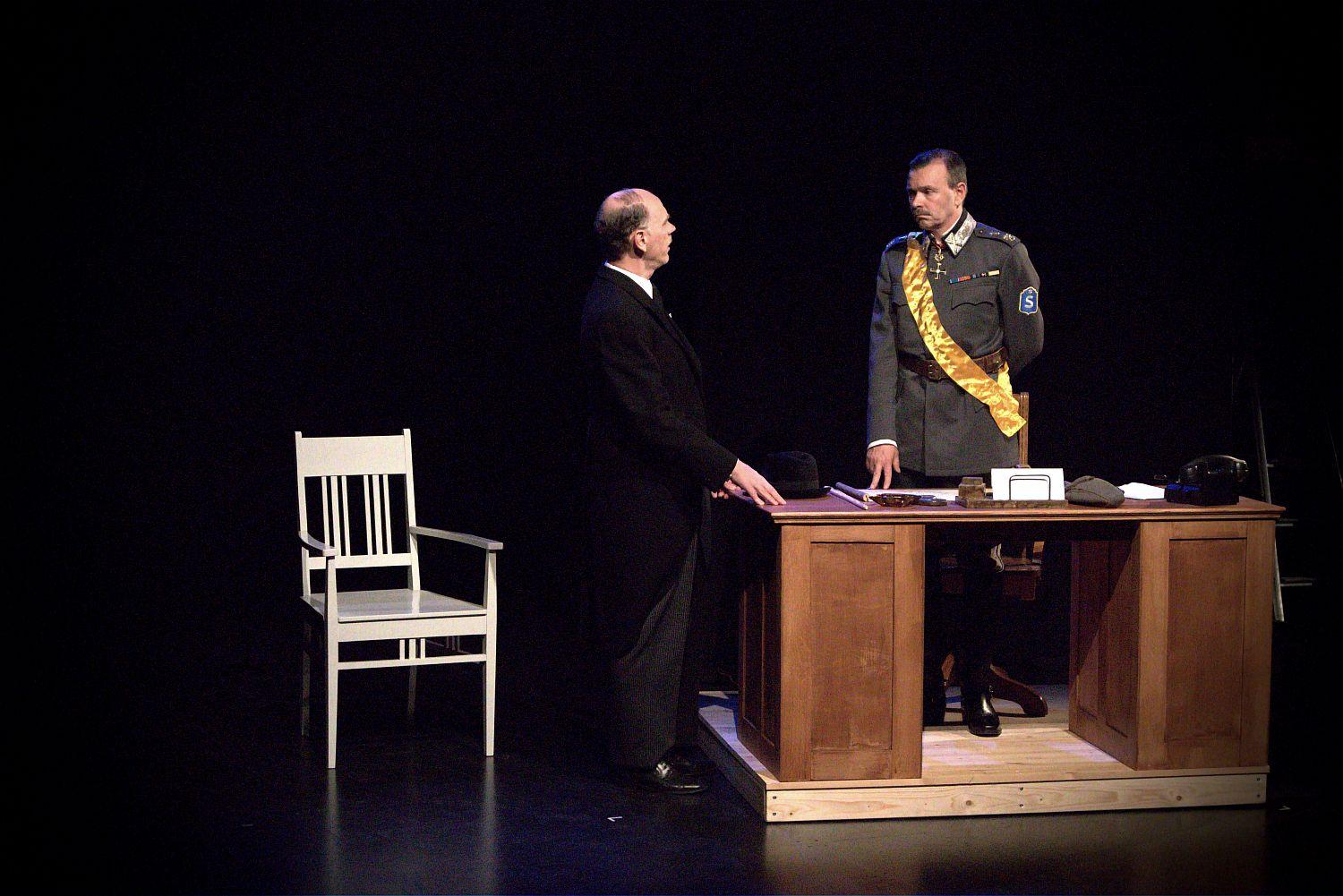En mörklagd teaterscen med marskalk Mannerheim bakom ett skrivbord, med en gäst iklädd jackett med ryggen mot kameran.
