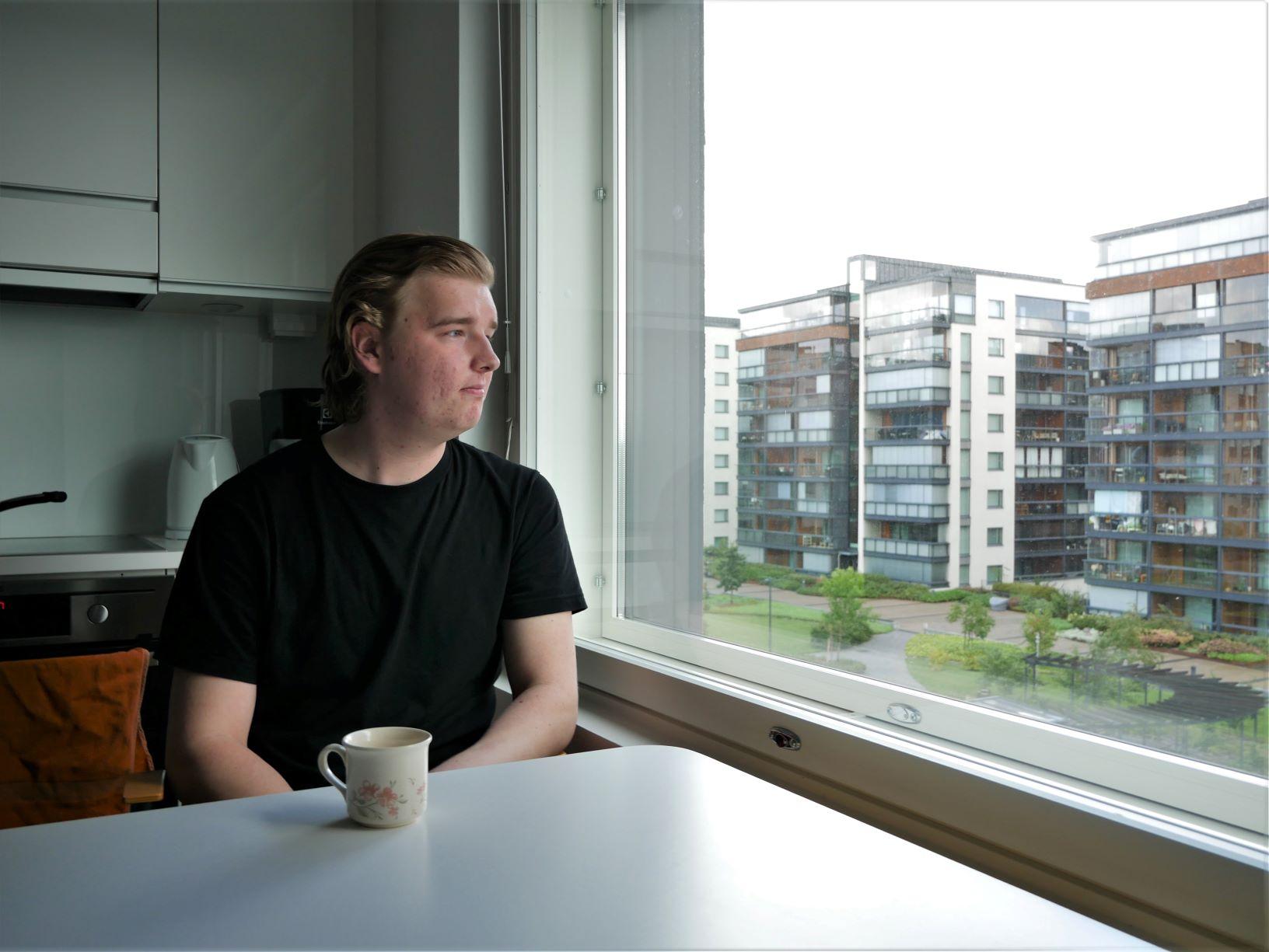 EN ung man sitter vid köksbordet och tittar ut genom fönstret. Det syns höghus i bakgrunden