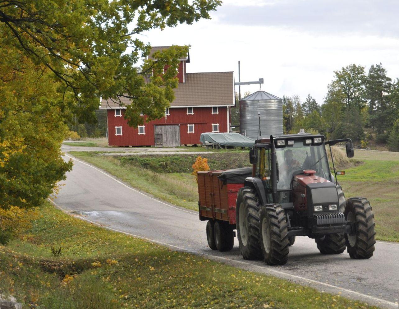 Traktor med släp kommer körande på vägen. I bakgrunden syns en tork.