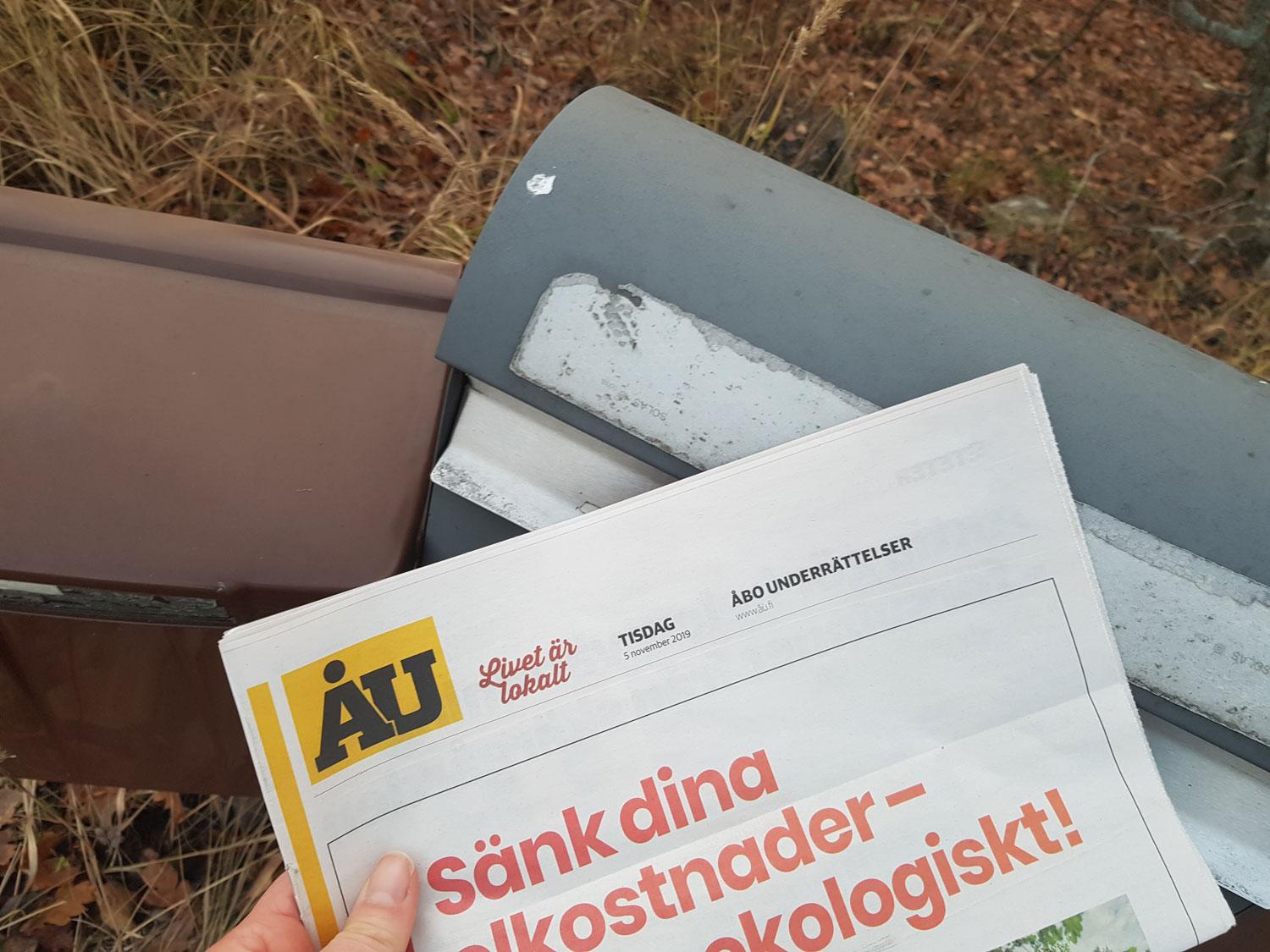 En hand håller en ÅU-tidning vid två postlådor