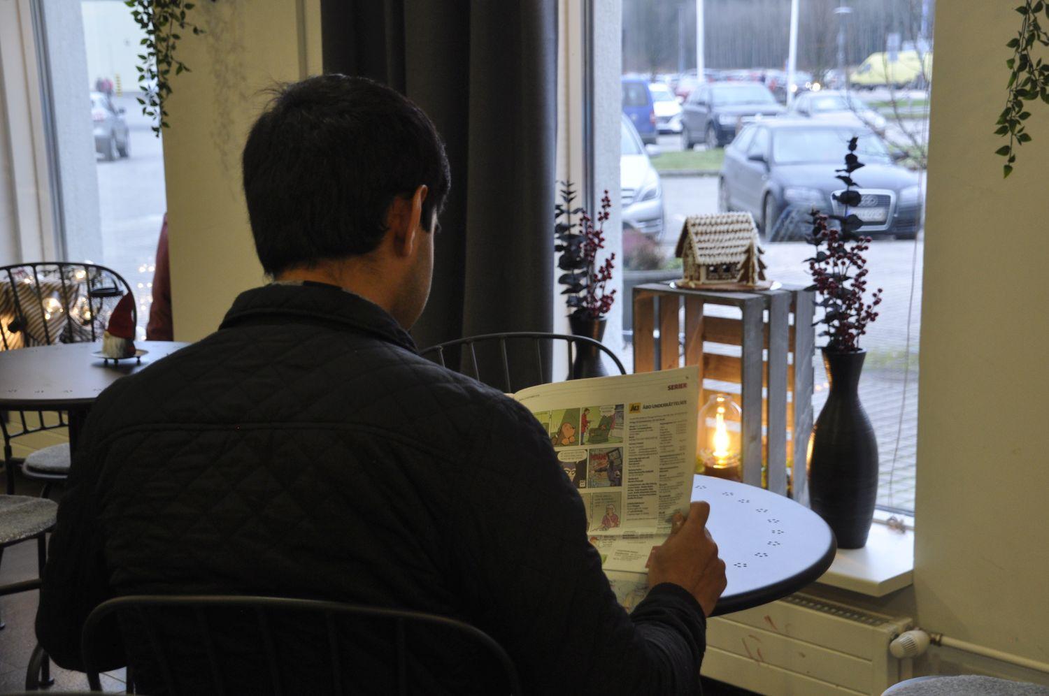 Man sitter vid ett cafébord och läser tidningen. Ett fönster med ett ljus synd framför mannen, som är fotad bakifrån.