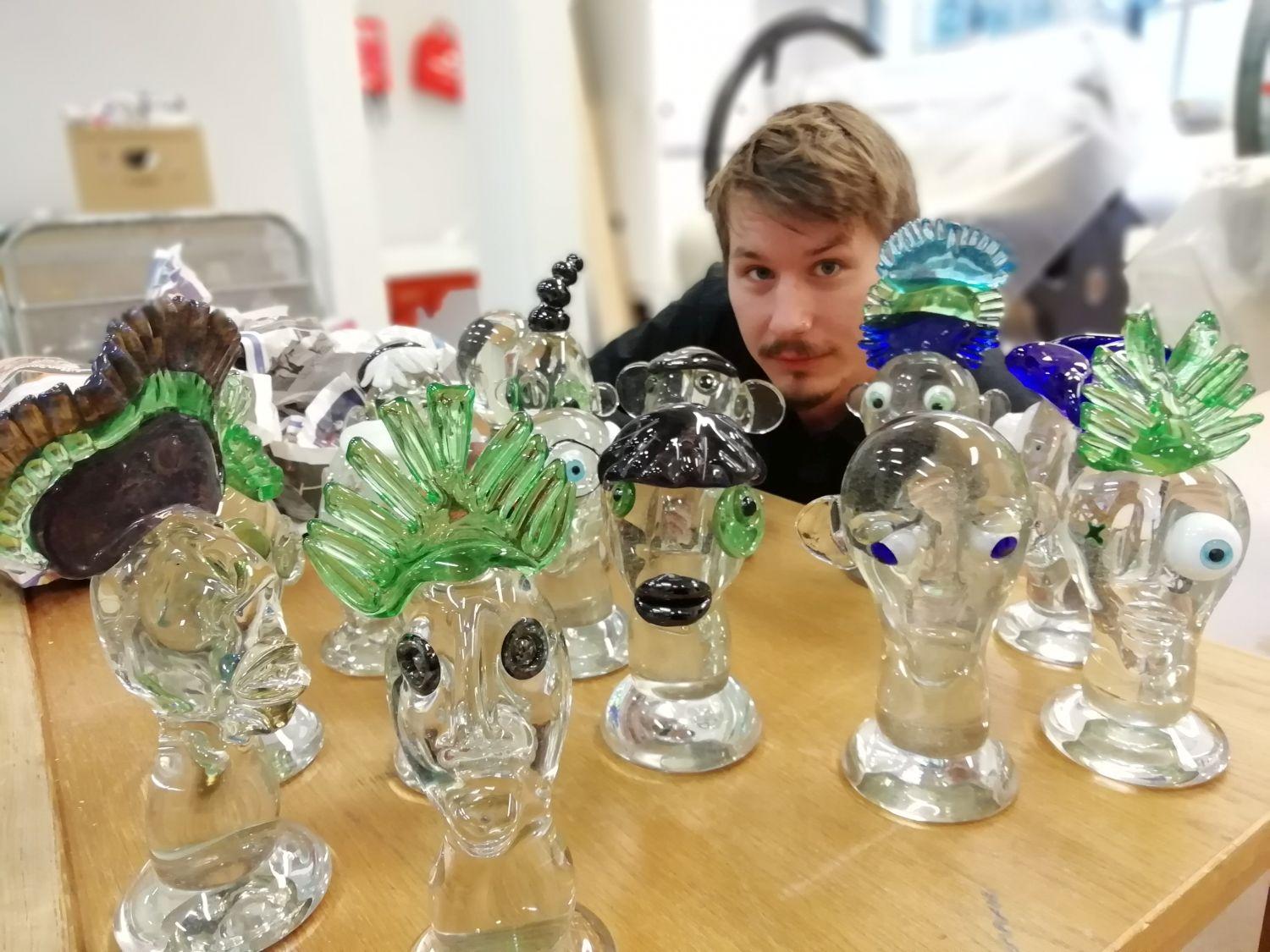 Glasskulpturer på ett bard. Bakom dem skymtar konstnären