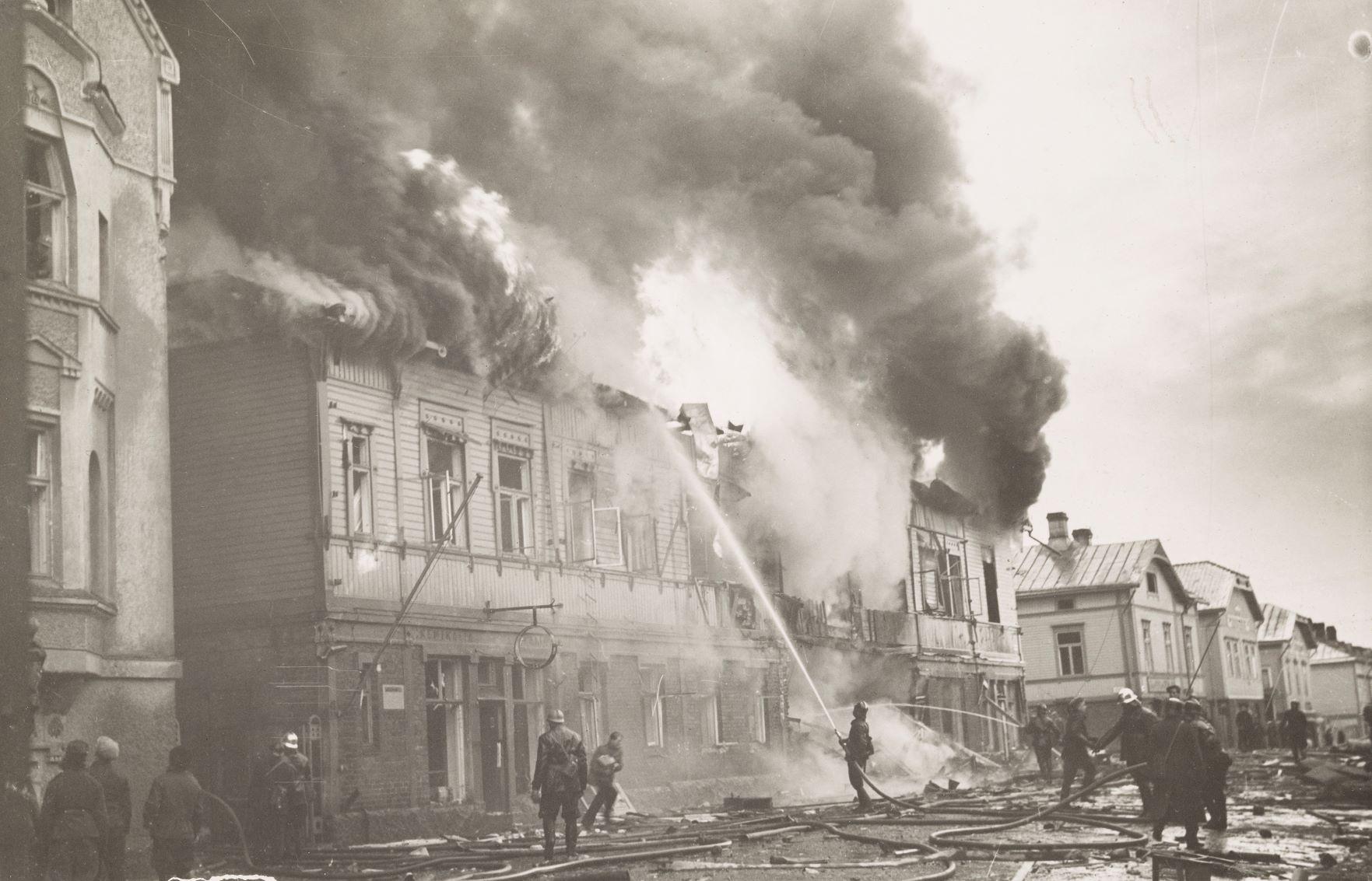 Ett svartvitt foto föreställandes ett trähus som brinner och brandkåren som försöker släcka
