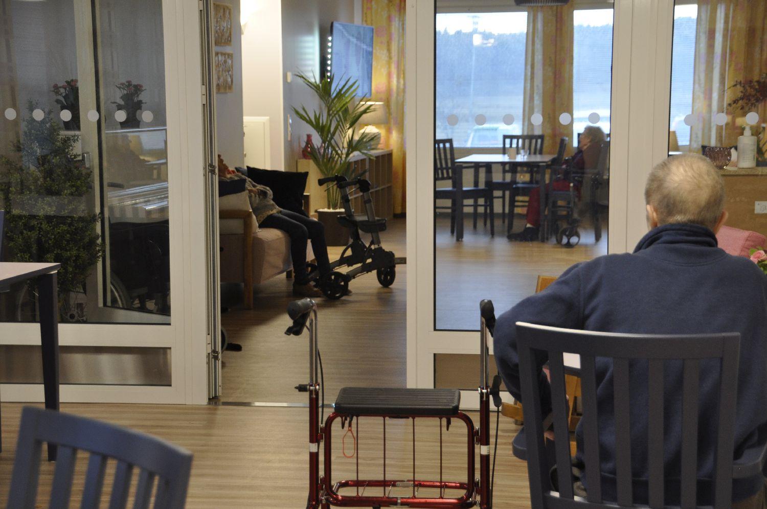 Två stora rum i ett äldreboende. Glasdörr skiljer dem åt. Äldre man och hans rollator syns bakifrån