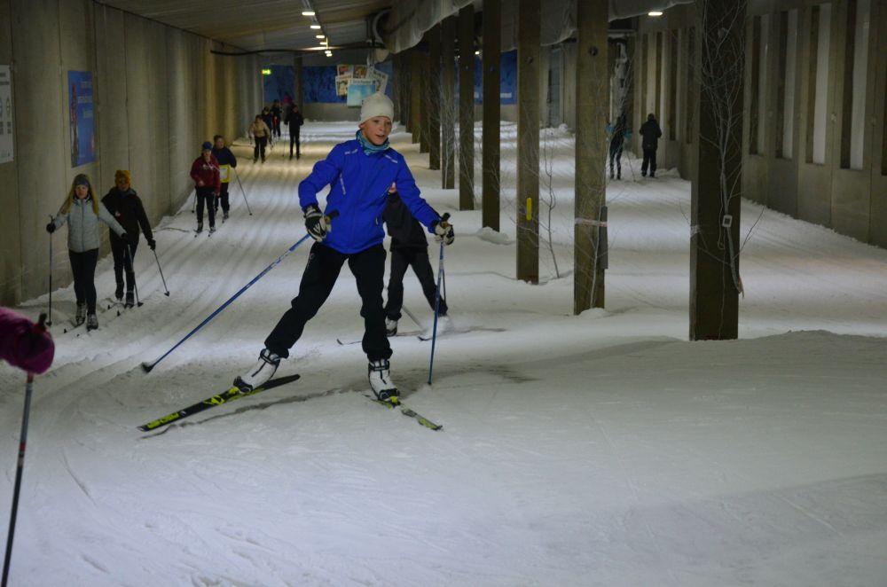 En vit pojke klädd i blått åker skidor längs ett inomhusskidspår