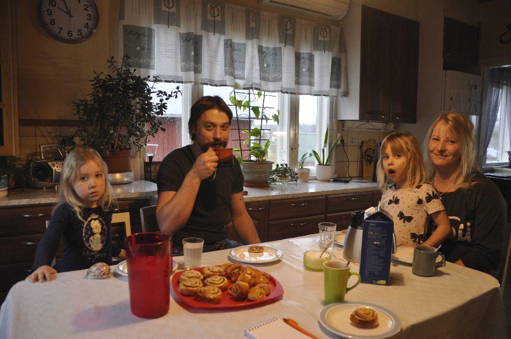 Familj dricker kaffe/saft och äter bulle i ett kök. Ett reporterblock syns på bordet