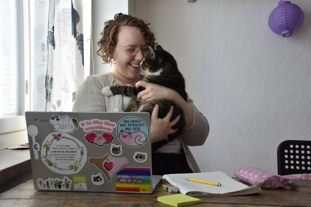 En ung kvinna sitter vid ett bord framför en dator och ler mot en katt hon håller i famnen.