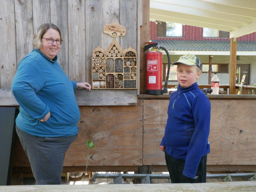 En mamma och ett barn står framför en trähylla som fyllts med träbitar det borrats hål i, metallgaller och kottar.