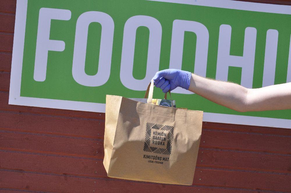 arm med en fylld kasse. Bakom syns en del av en grön skylt med texten foodhub.