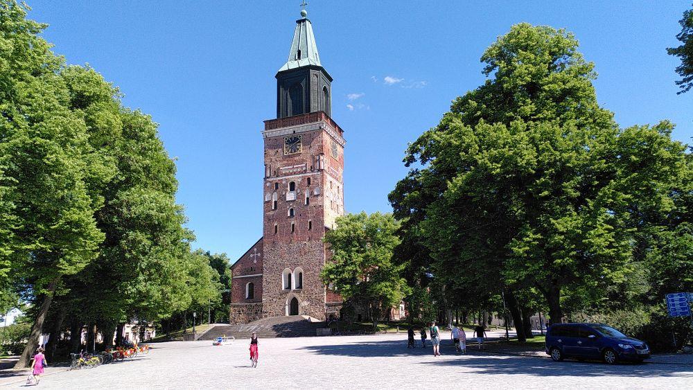 Ett kyrkotorn omringat av gröna träd. Framför kyrkan går och cyklar människor.