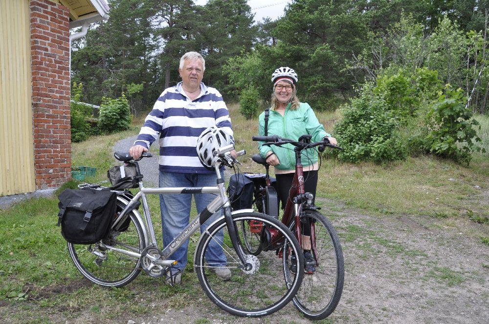 Två personer, den ena med cykelhjälm, står vid sina cyklar. Bakom dem natur och hörnet av en tegelbyggnad.