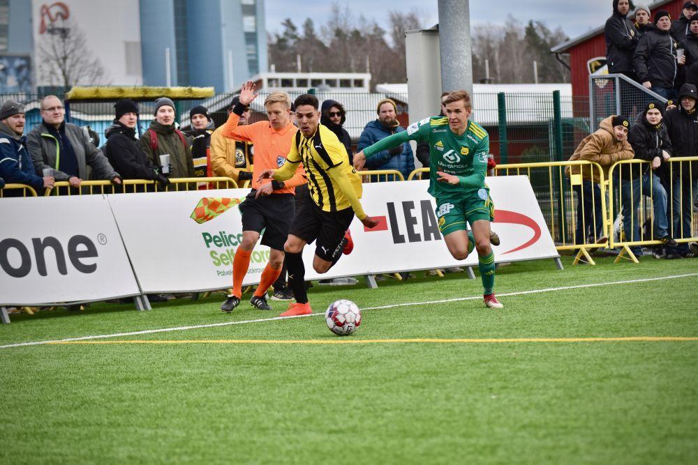 Två manliga fotbollsspelare, en i gul tröja och en i grön, jagar en boll på en fotbollsplan.
