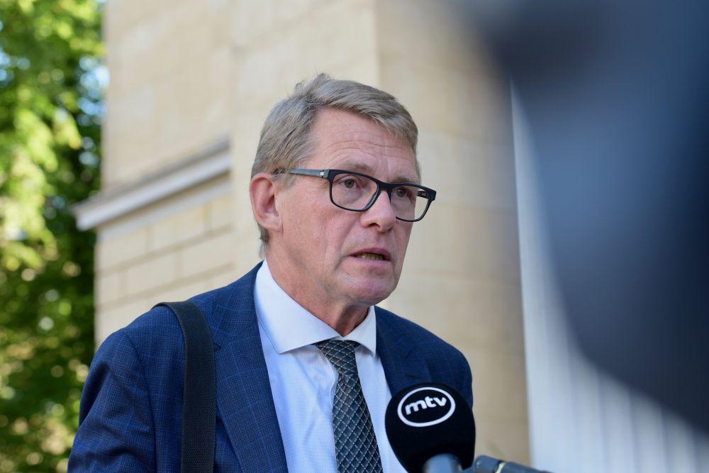 Kostymklädd äldre man står utomhus och pratar i en mikrofon.